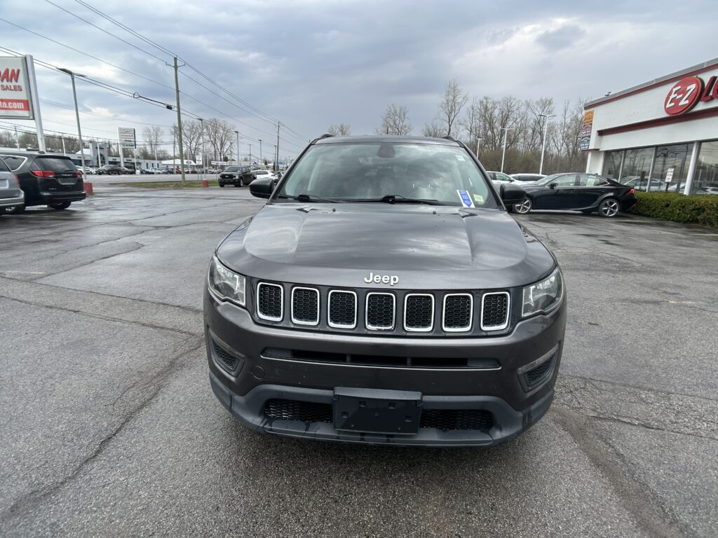 Front view of a gray Jeep SUV parked in a car dealership lot with other cars and a building in the background.