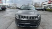 Front view of a gray Jeep SUV parked in a car dealership lot with other cars and a building in the background.