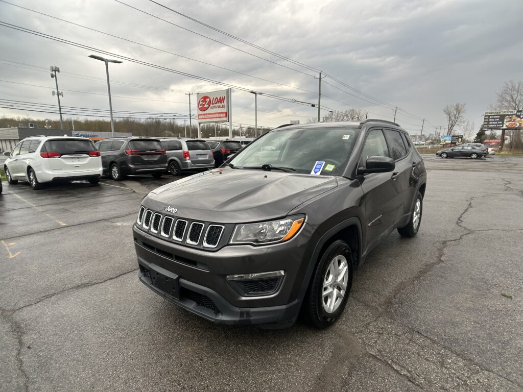 Gray Jeep SUV in foreground at a car dealership lot with a row of parked cars and an EZ Loan sign in the background under a cloudy sky, the scene wide and open.