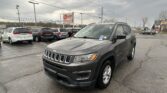 Gray Jeep SUV in foreground at a car dealership lot with a row of parked cars and an EZ Loan sign in the background under a cloudy sky, the scene wide and open.