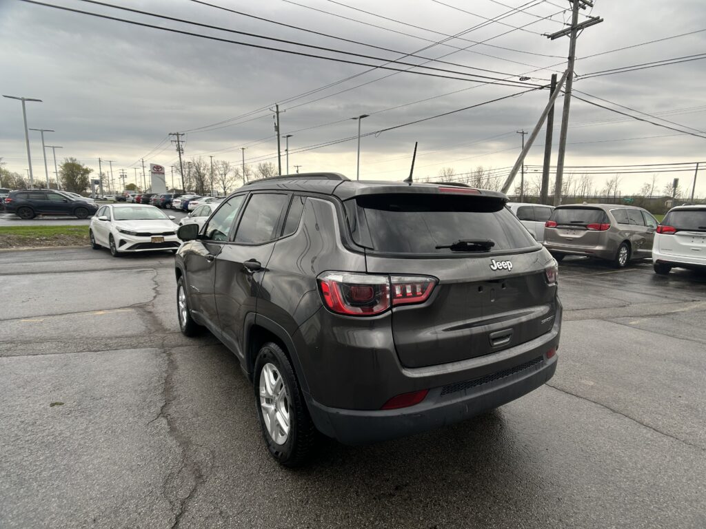 Rear view of a gray Jeep SUV in a dealership parking lot with other cars and overcast sky above.