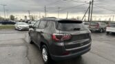 Rear view of a gray Jeep SUV in a dealership parking lot with other cars and overcast sky above.