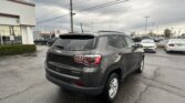 Rear three-quarter view of a dark gray Jeep SUV parked in a car dealership lot, with a Toyota dealership building in the background and other cars lined up nearby.