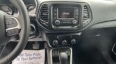 Inside a Jeep cabin showing the steering wheel on the left, center touchscreen with 'Jeep', climate controls, gear shifter, and a paper mat reading 'We Take Pride In Your Vehicle'.