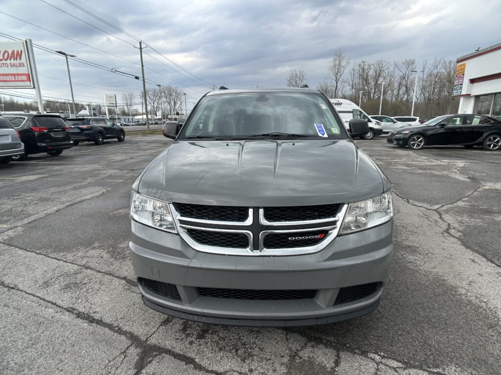 Front view of a gray Dodge SUV in a car dealership lot, with other cars and a large sign in the background, under a cloudy sky.