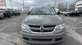 Front view of a gray Dodge SUV in a car dealership lot, with other cars and a large sign in the background, under a cloudy sky.