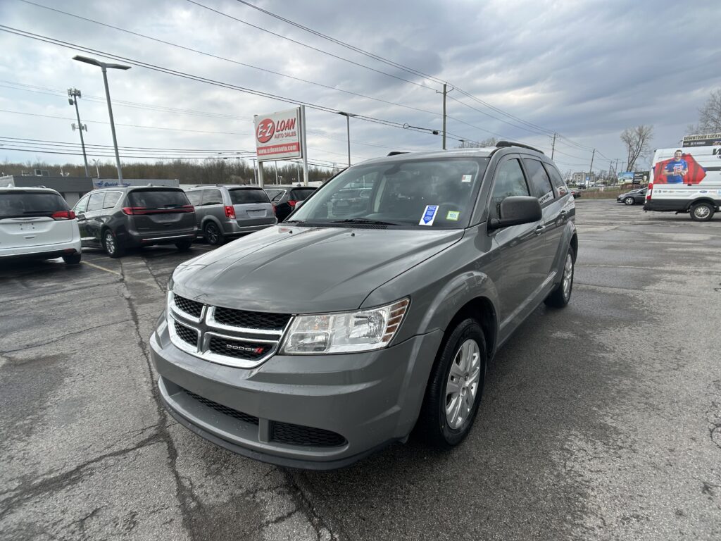 Front-right view of a gray Dodge minivan in a car dealership lot, with other vehicles and an EZ LOAN Auto Sales sign in the background under cloudy skies.