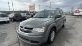 Front-right view of a gray Dodge minivan in a car dealership lot, with other vehicles and an EZ LOAN Auto Sales sign in the background under cloudy skies.