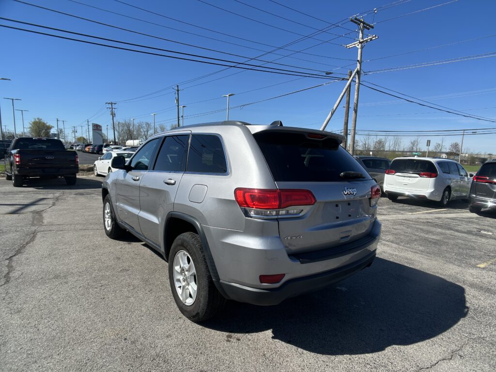 Rear three-quarter view of a silver Jeep Grand Cherokee in a crowded parking lot under a clear blue sky with overhead power lines and other cars nearby.