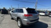 Rear three-quarter view of a silver Jeep Grand Cherokee in a crowded parking lot under a clear blue sky with overhead power lines and other cars nearby.
