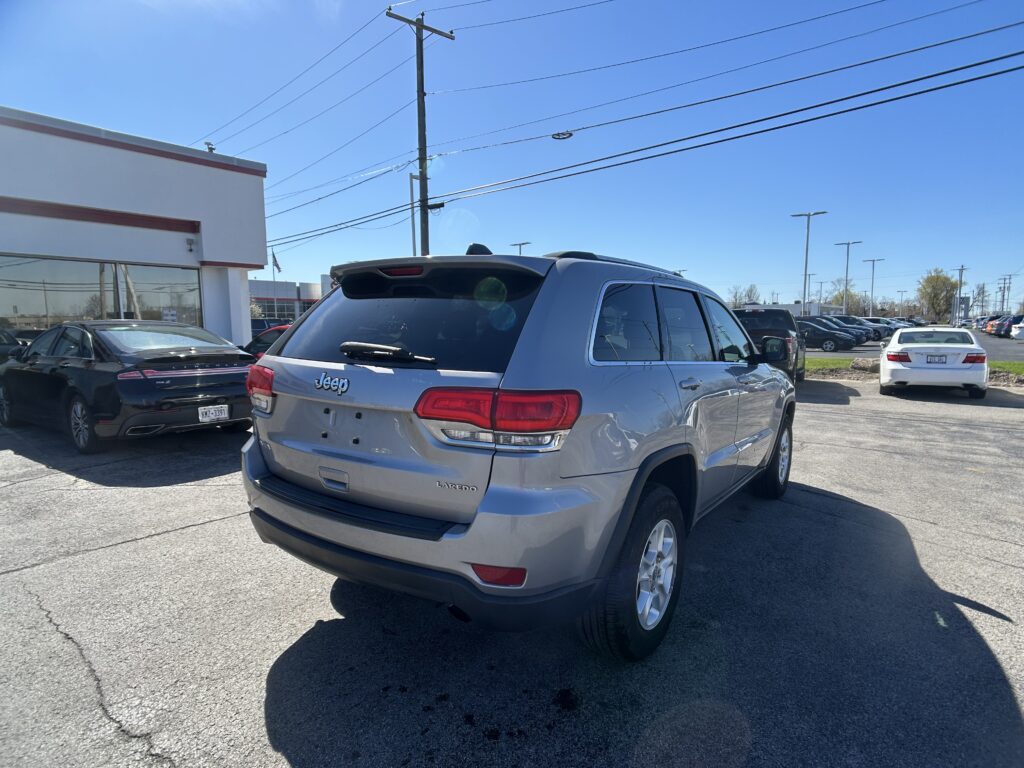 Rear of a silver Jeep Grand Cherokee Laredo in a car dealership lot, with a white building on the left and multiple cars parked nearby under a clear blue sky.