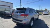 Rear of a silver Jeep Grand Cherokee Laredo in a car dealership lot, with a white building on the left and multiple cars parked nearby under a clear blue sky.