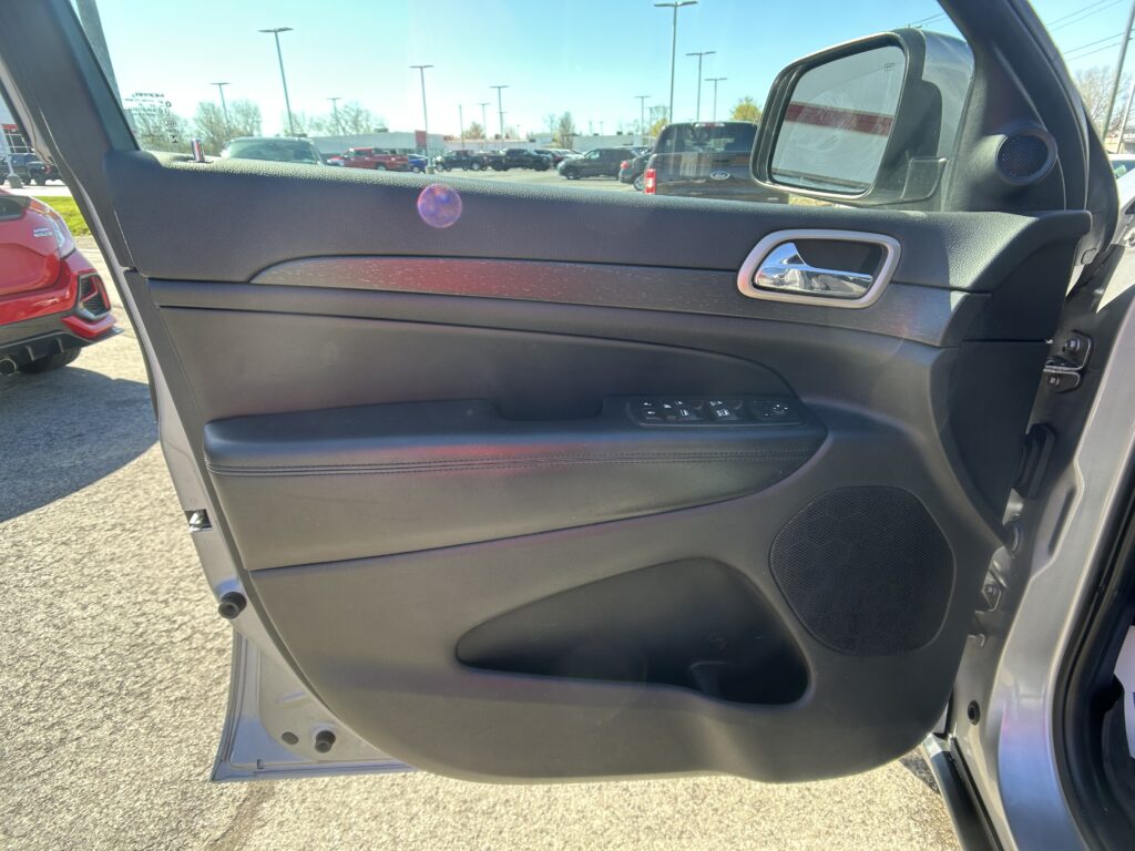 Interior of a car door with window controls and chrome handle; parking lot visible through the open door on a sunny day