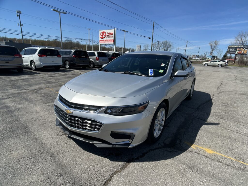 Silver Chevrolet sedan in the foreground of a car dealership lot with multiple minivans parked behind and an EZ Loan Auto Sales sign in the background under a clear blue sky.