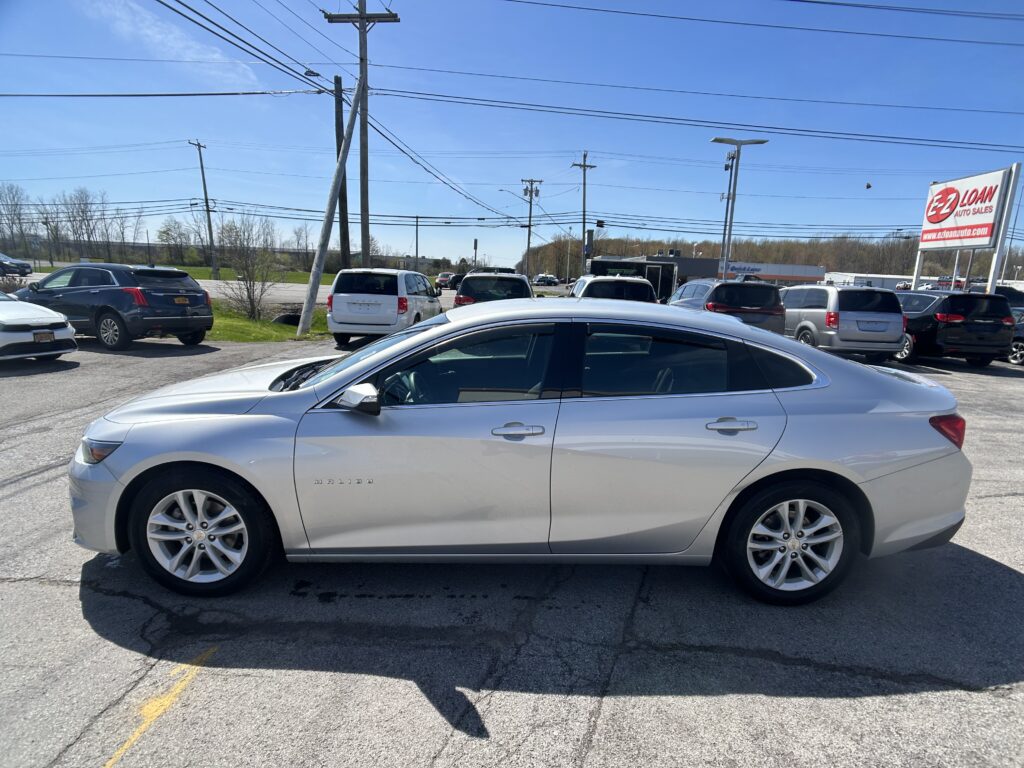 Silver Chevy Malibu in a car dealership lot with other SUVs, power lines overhead, and a tall EZ LOAN Auto Sales sign to the right under a blue sky.