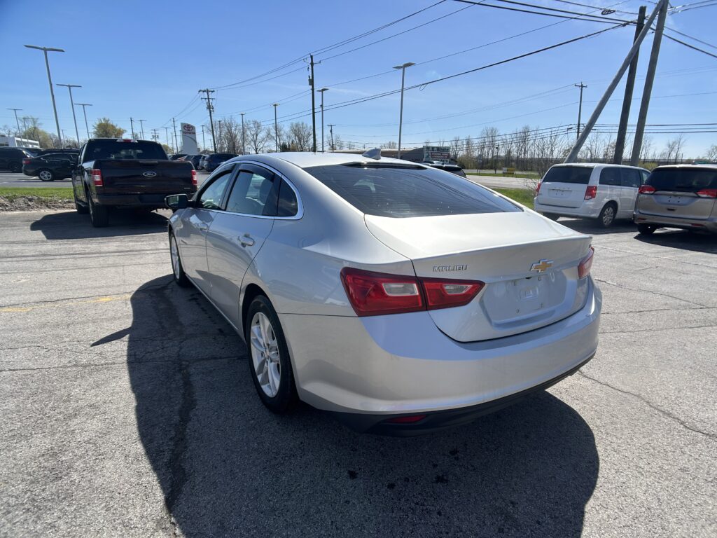 Silver Chevy Malibu in the foreground parking lot with multiple parked cars and tall power lines overhead on a sunny day.
