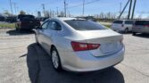 Silver Chevy Malibu in the foreground parking lot with multiple parked cars and tall power lines overhead on a sunny day.