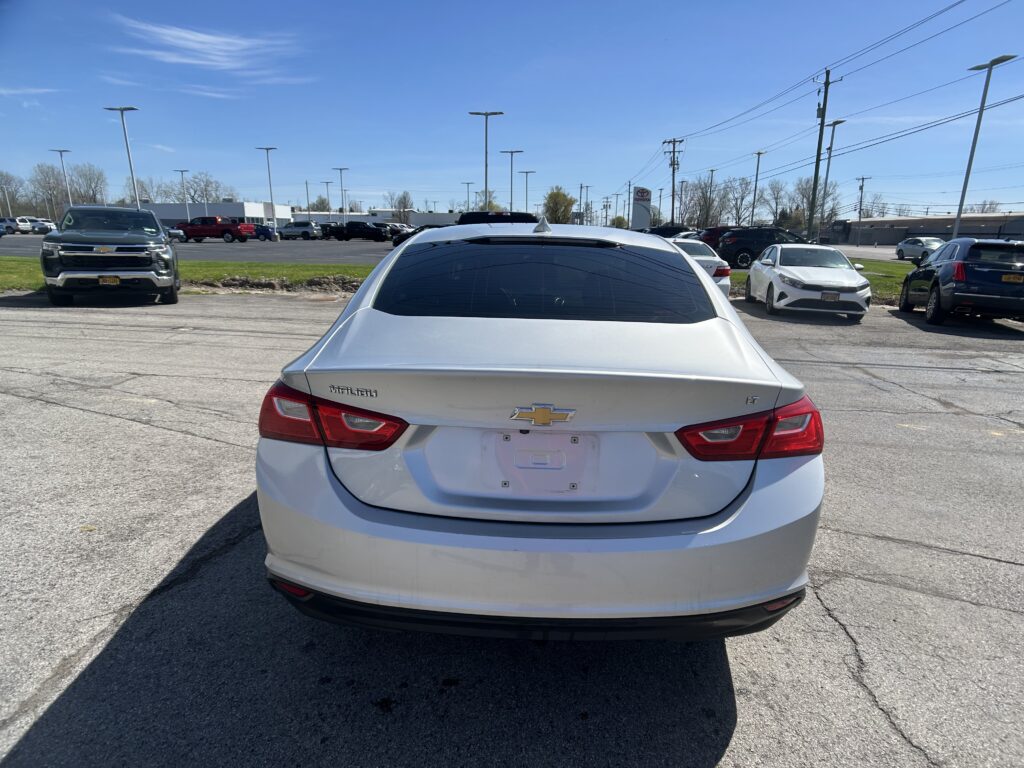 Rear view of a white Chevrolet Malibu in a dealership parking lot with other cars and a clear blue sky behind it.