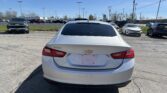 Rear view of a white Chevrolet Malibu in a dealership parking lot with other cars and a clear blue sky behind it.
