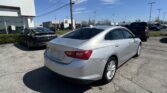 Silver sedan in the foreground at a Toyota dealership lot, with a white building on the left and a clear blue sky behind.