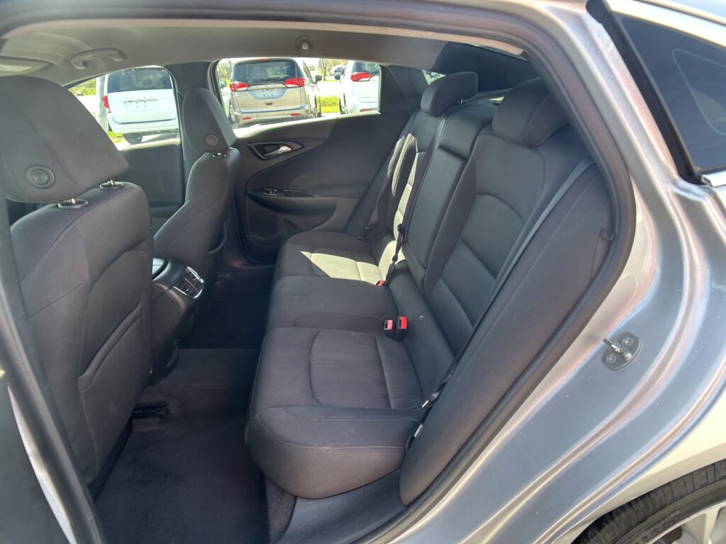 Rear interior of a dark cloth car with three headrests and seatbelts, door panel visible on the left, parked cars outside through the window.
