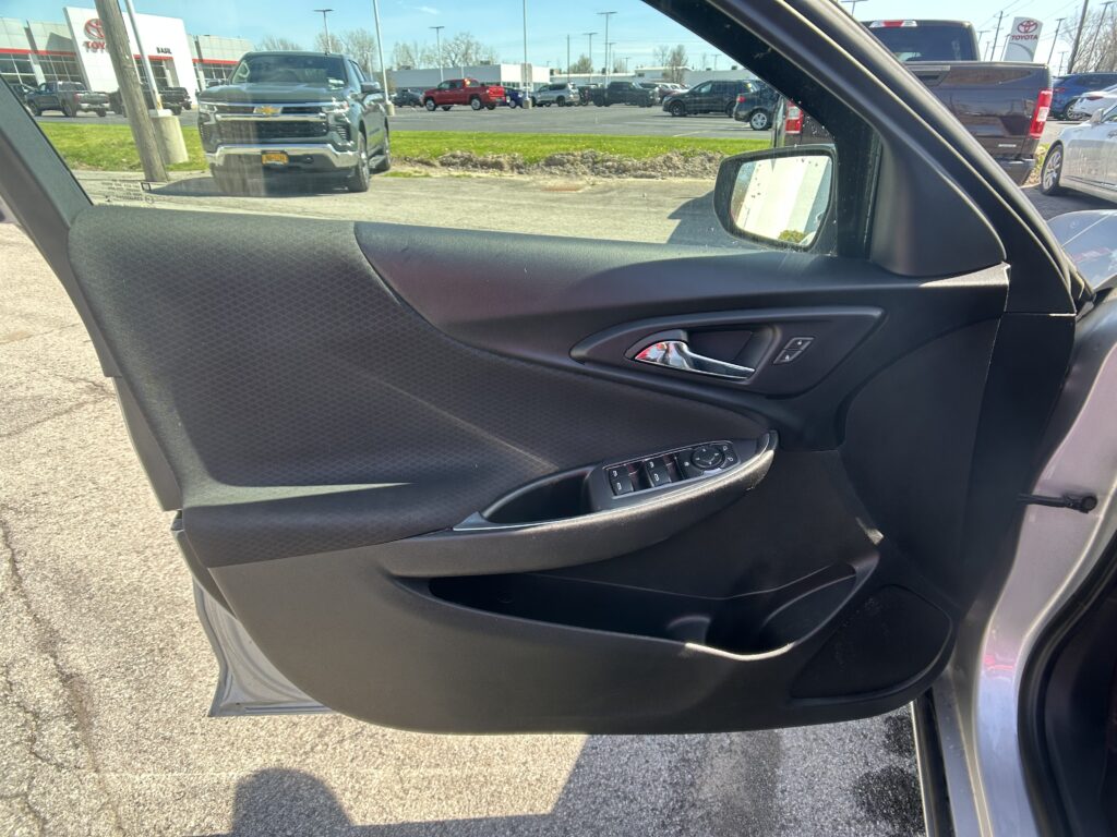Close-up of a black car door panel with window controls, viewed from inside; outside is a dealership lot with trucks and cars in the background.
