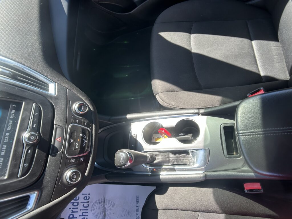 Interior of a car showing the center console with controls, gear shifter, and cup holder area; passenger seat and dashboard visible with sunlight casting shadows on the seat.