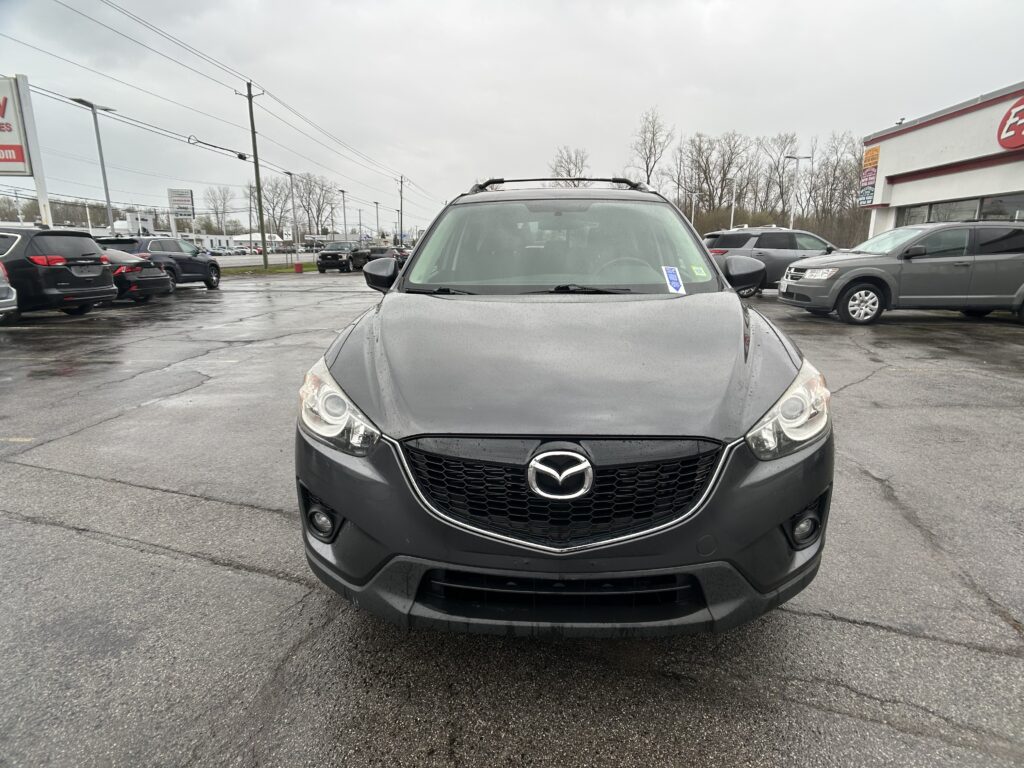 Front view of a dark gray Mazda SUV with a roof rack parked in a wet dealership lot on a cloudy day.