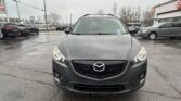 Front view of a dark gray Mazda SUV with a roof rack parked in a wet dealership lot on a cloudy day.