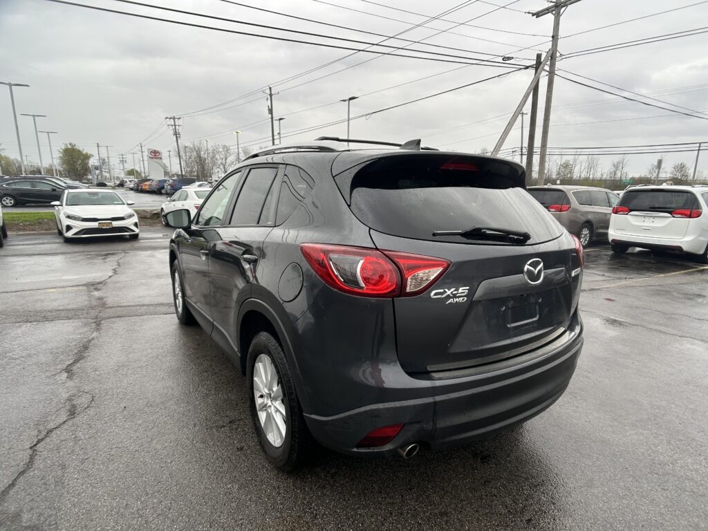Rear three-quarter view of a gray Mazda CX-5 AWD in a dealership parking lot, with other cars and overhead power lines in the background on a damp day.