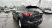 Rear three-quarter view of a gray Mazda CX-5 AWD in a dealership parking lot, with other cars and overhead power lines in the background on a damp day.