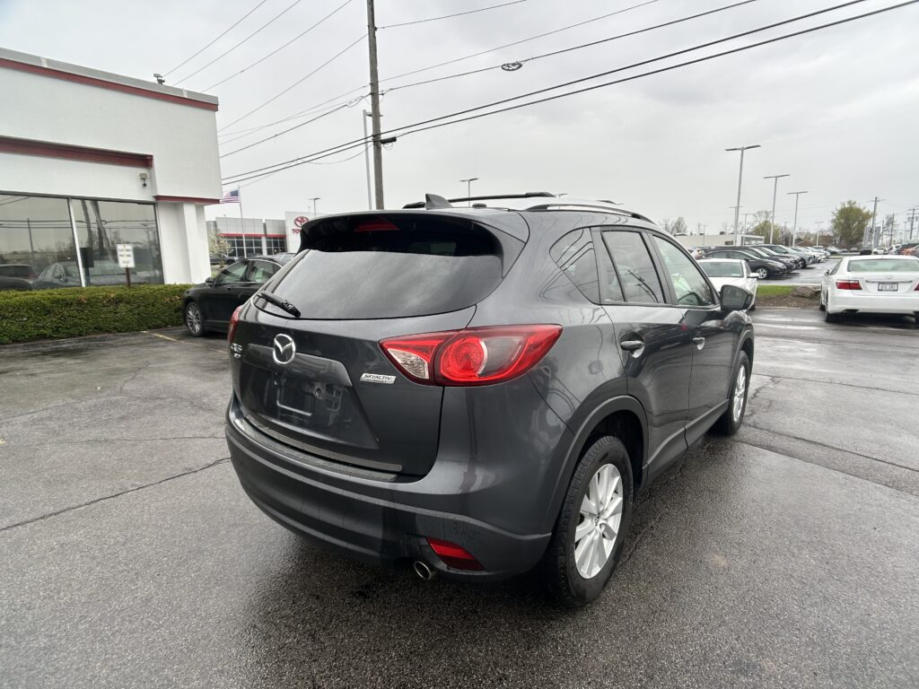 Rear three-quarter view of a gray Mazda SUV in a dealership lot, with a showroom building on the left and other cars in the background.