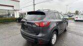 Rear three-quarter view of a gray Mazda SUV in a dealership lot, with a showroom building on the left and other cars in the background.