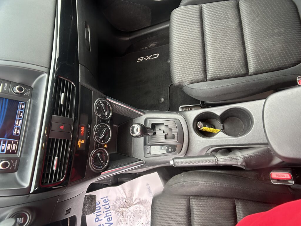 Interior of a dark SUV showing the center console with gear shifter, climate controls, cup holders, and a CX-5 branded floor mat near the driver seat with black fabric seats and side dash visible.