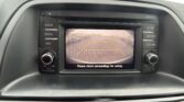 Car infotainment screen showing rearview camera with yellow parking lines and 'Please check surroundings for safety' text visible.