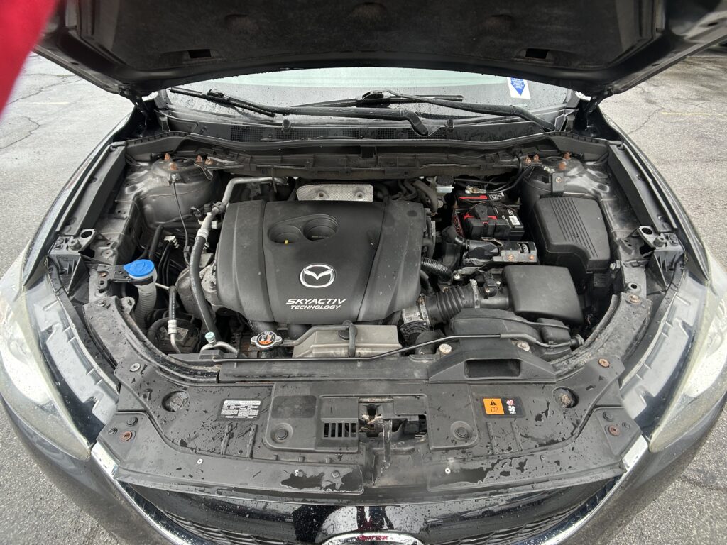 Car hood open, engine bay visible with Mazda SkyActiv Technology cover and battery on the right.