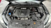 Car hood open, engine bay visible with Mazda SkyActiv Technology cover and battery on the right.