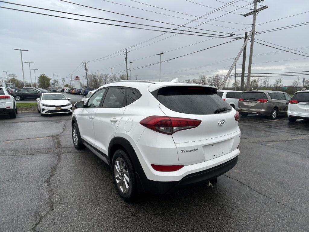 White Hyundai Tucson in the foreground of a crowded car dealership lot on a rainy day, with many cars parked and power lines overhead.