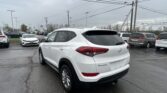 White Hyundai Tucson in the foreground of a crowded car dealership lot on a rainy day, with many cars parked and power lines overhead.