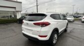 White Hyundai SUV photographed from the rear three-quarters, parked in a dealership lot with other cars and a white building in the background on a cloudy day.
