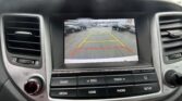 Car infotainment screen showing a rearview camera view with parking guidelines: yellow guide lines and a red line, surrounding cars visible in a parking lot, text reads 'Check surroundings for your safety.'