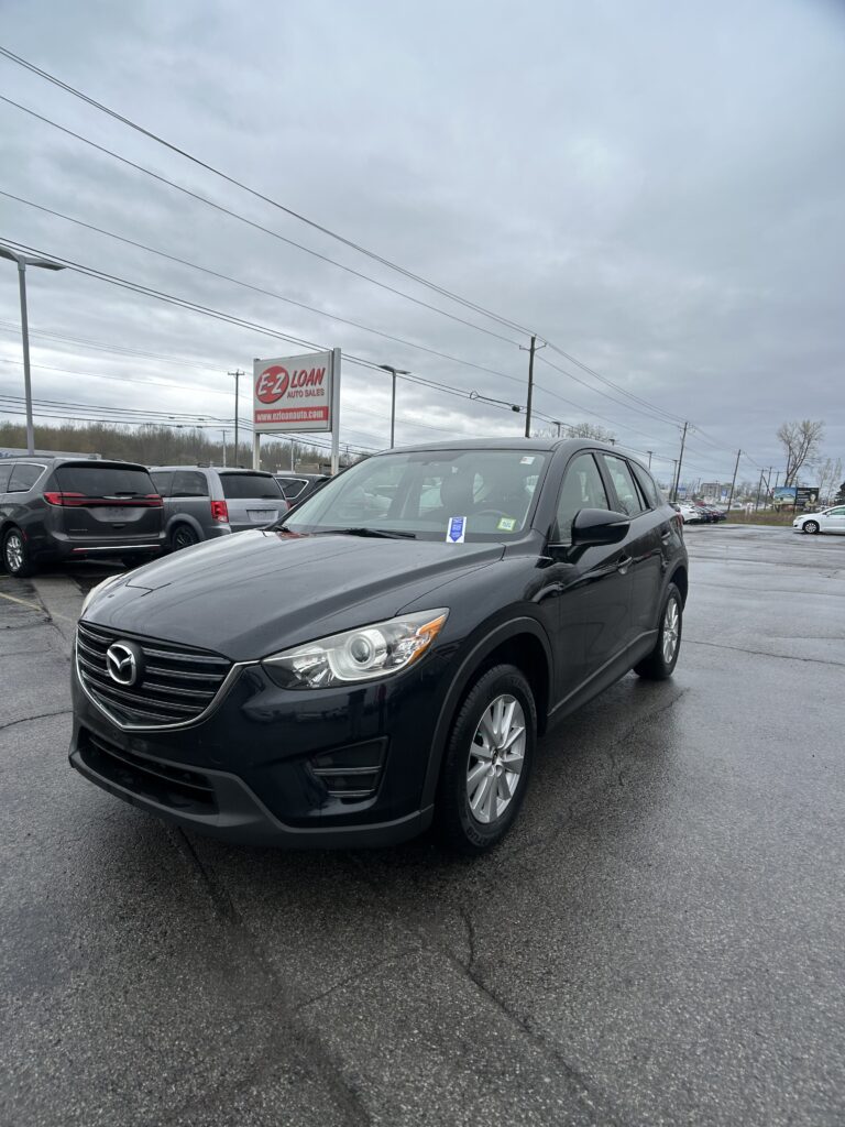 Dark gray Mazda SUV in a car dealership lot, with an E-Z Loan Auto Sales sign in the background and a cloudy sky above.