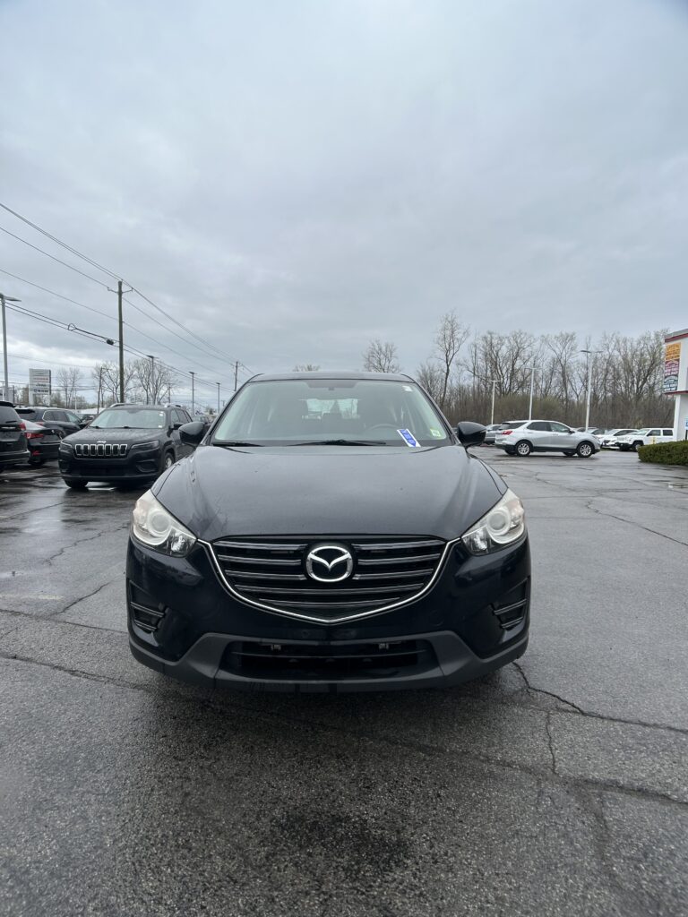 Front view of a black Mazda SUV in a car dealership lot, with other cars and a cloudy sky in the background.