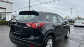 Rear three-quarter view of a black Mazda SUV parked in a dealership lot on a cloudy day, with other cars in the background and a building to the left.