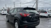 Rear three-quarter view of a black Mazda CX-5 in a car dealership lot, with white cars and power lines overhead on a cloudy day.