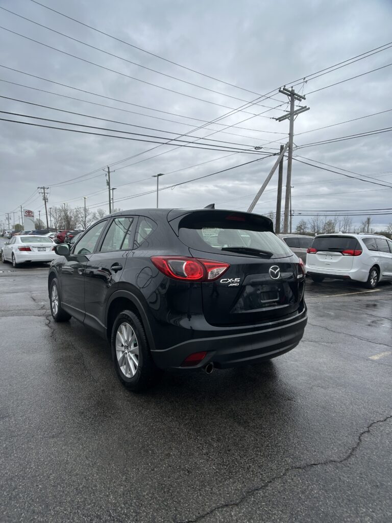 Rear three-quarter view of a black Mazda CX-5 in a car dealership lot, with white cars and power lines overhead on a cloudy day.