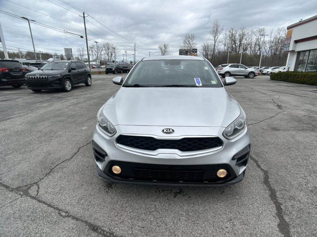 Front view of a silver Kia SUV in a car dealership parking lot with other vehicles and a building in the background under cloudy skies.