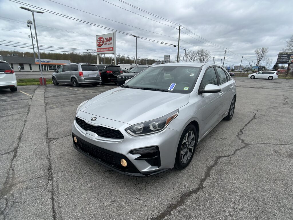 Silver sedan (Kia) in the foreground of a car lot, with other vehicles and an EZ LOAN Auto Sales sign in the background on a cloudy day.