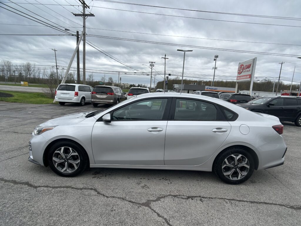 Silver sedan in the foreground at a car dealership lot with other minivans and SUVs parked behind it under a cloudy sky and power lines overhead, EZ LOAN Auto Sales sign visible in the distance.
