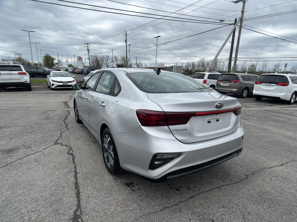 Rear view of a silver Kia Forte in a busy car dealership lot on a cloudy day while other vehicles surround it.